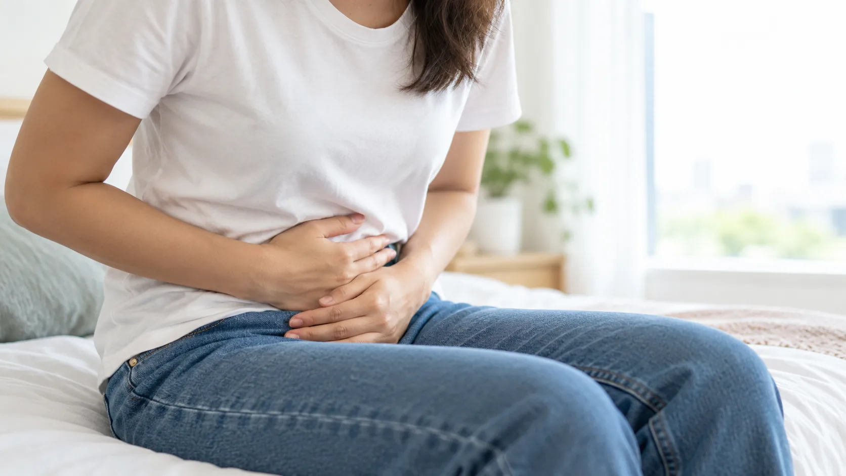 Woman sitting on a bed holding her lower abdomen, experiencing pelvic pain