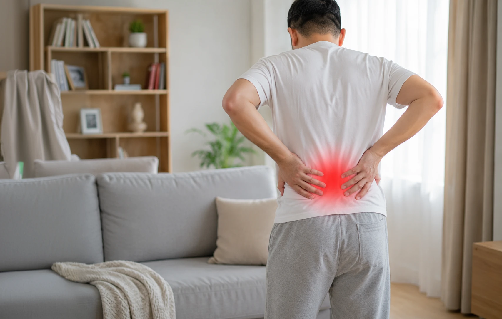 Man standing at home with both hands on his lower back, representing lower back pain or back pain symptoms.